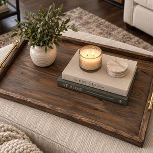 Peut inclure: Un plateau de service rectangulaire en bois brun foncé avec deux poignées en métal doré. Le plateau contient un petit vase en céramique blanche avec du feuillage vert, une bougie allumée, deux livres intitulés The Kinfolk Home et At Home, et des sous-verres.
