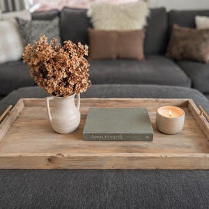 May include: A wooden tray with brass handles displays a white ceramic jug with dried flowers, a green book with the text "down to earth", and a lit candle in a beige holder. The tray is on a dark grey ottoman in front of a grey sofa.