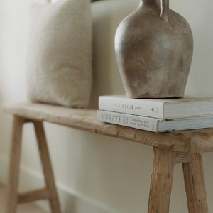 Puede incluir: Un banco de madera rústica con un acabado claro. Un cojín texturizado de color crema descansa sobre el banco. Un jarrón gris con asa contiene ramas. Dos libros están apilados en el banco.