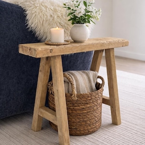 May include: A rustic wooden bench with a candle, vase of white flowers, and a woven basket. The bench has a natural wood finish and a simple A-frame design. The basket contains a folded blanket.