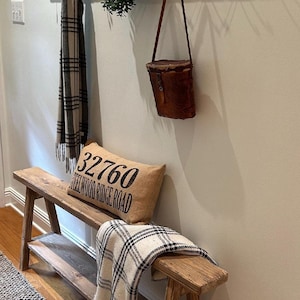 May include: A rustic entryway scene featuring a wooden bench with a shelf, a plaid throw, and a burlap pillow with text. A wooden coat rack with hooks holds a scarf and a leather bag. Decorative elements include a woven wall hanging and greenery.