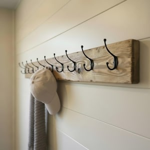 May include: A rustic wooden coat rack with ten black metal hooks. A light-colored baseball cap and a gray scarf hang from the hooks. The rack is mounted on a cream-colored wall with horizontal paneling.