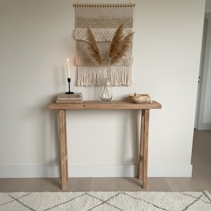 Puede incluir: Una consola de madera de color claro con un tapiz de pared tejido, un jarrón con plantas secas, una vela y un cuenco de madera pequeño. La mesa está contra una pared blanca y tiene una alfombra delante.