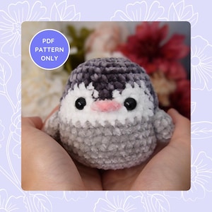 Puede incluir: Un juguete de amigurumi de pingüino tejido a crochet con pelaje gris y blanco, ojos negros y un pico rosa. El juguete está siendo sostenido en las manos de una persona.