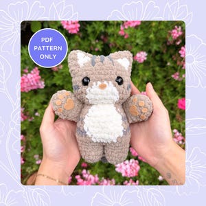 Può includere: Un peluche a forma di gatto all'uncinetto nei toni del marrone, grigio e bianco, tenuto in mano. Il gatto ha un motivo a strisce, occhi neri e dettagli delle zampe. Una grafica circolare in alto a sinistra recita "PDF PATTERN ONLY".