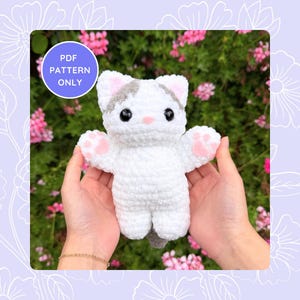 Peut inclure: Un chat blanc au crochet avec des détails gris et roses, notamment des taches grises sur la tête et des pattes roses. Le chat a des yeux de sécurité noirs et un nez rose. Le texte "PDF PATTERN ONLY" est inclus.