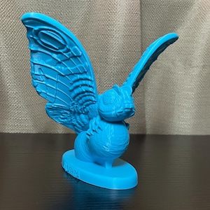 May include: A vibrant, light blue moth figurine with detailed wing patterns and a rounded body. The sculpture is mounted on a rectangular base, showcasing a textured surface. The moth's wings are spread, giving the impression of flight.