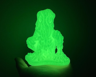 May include: A glowing green, semi-translucent figurine of a creature with a textured, organic surface. The figure features a prominent head with defined facial features and raised arms, standing on a flat base with text embossed on the front.