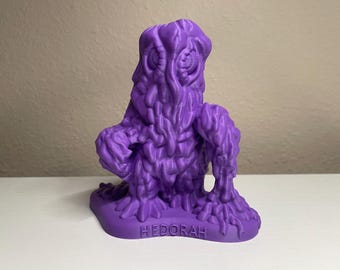 May include: A purple 3D printed figurine of the creature Hedorah, featuring a textured, sludge-like body with visible layered details, large circular eyes, and the name HEDORAH embossed on the front of the base.