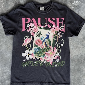 May include: A black short-sleeve t-shirt featuring a graphic print with the text PAUSE in pink and NEVER MIND in green. The design includes a mahjong tile with a peacock, pink lotus flowers, a butterfly, and white cherry blossom accents.