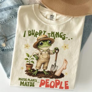 Może przedstawiać: Kremowy t-shirt z grafiką zielonej żaby w słomkowym kapeluszu i ogrodniczkach, trzymającej łopatę w ziemi. Napis głosi I BURRY THINGS... MAYBE PLANTS... MAYBE PEOPLE. Wokół żaby znajdują się małe żółte kwiatki.