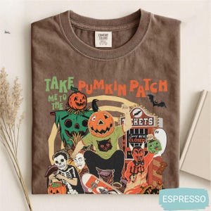 Puede incluir: Una camiseta marrón con un estampado gráfico que incluye el texto "TAKE PUMPKIN PATCH ME TO THE". El diseño muestra una figura con cabeza de calabaza en un monopatín, un espantapájaros, un esqueleto y un diablo frente a una taquilla.