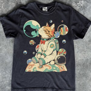 May include: A black short-sleeved t-shirt featuring a graphic of a cat wearing a space suit and a red bow tie, sitting on a planet. The cat is surrounded by colourful floating orbs and planets in space, with a detached space helmet resting nearby.