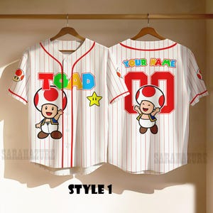 May include: White baseball jersey with red pinstripes, featuring a cartoon Toad character and the word "TOAD" in colorful letters. The back displays "YOUR NAME" and the number "00".