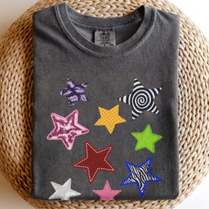 Peut inclure: Un t-shirt à col rond gris anthracite orné de plusieurs étoiles colorées. Les étoiles présentent divers motifs, notamment un imprimé marbré violet, une texture à sequins roses, une spirale noire et blanche, des pois jaunes, du vert lime, du rouge, du blanc, du rose uni et un motif zébré bleu et blanc.
