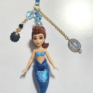 May include: A mermaid doll keychain with a blue tail and top. The doll has brown hair and is attached to a blue heart-shaped clasp. Decorative beads and a seashell charm dangle from the chain.