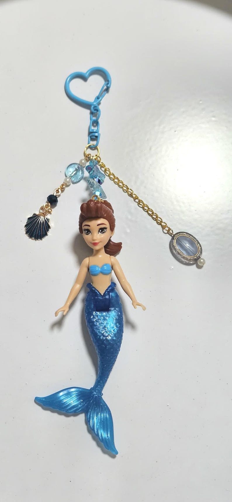 May include: A mermaid doll keychain with a blue tail and top. The doll has brown hair and is attached to a blue heart-shaped clasp. Decorative beads and a seashell charm dangle from the chain.