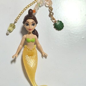 May include: A mermaid keychain featuring a yellow tail and green top. The doll has brown hair and is attached to a gold chain with decorative beads and a yellow heart-shaped clasp. A green seashell charm is also included.