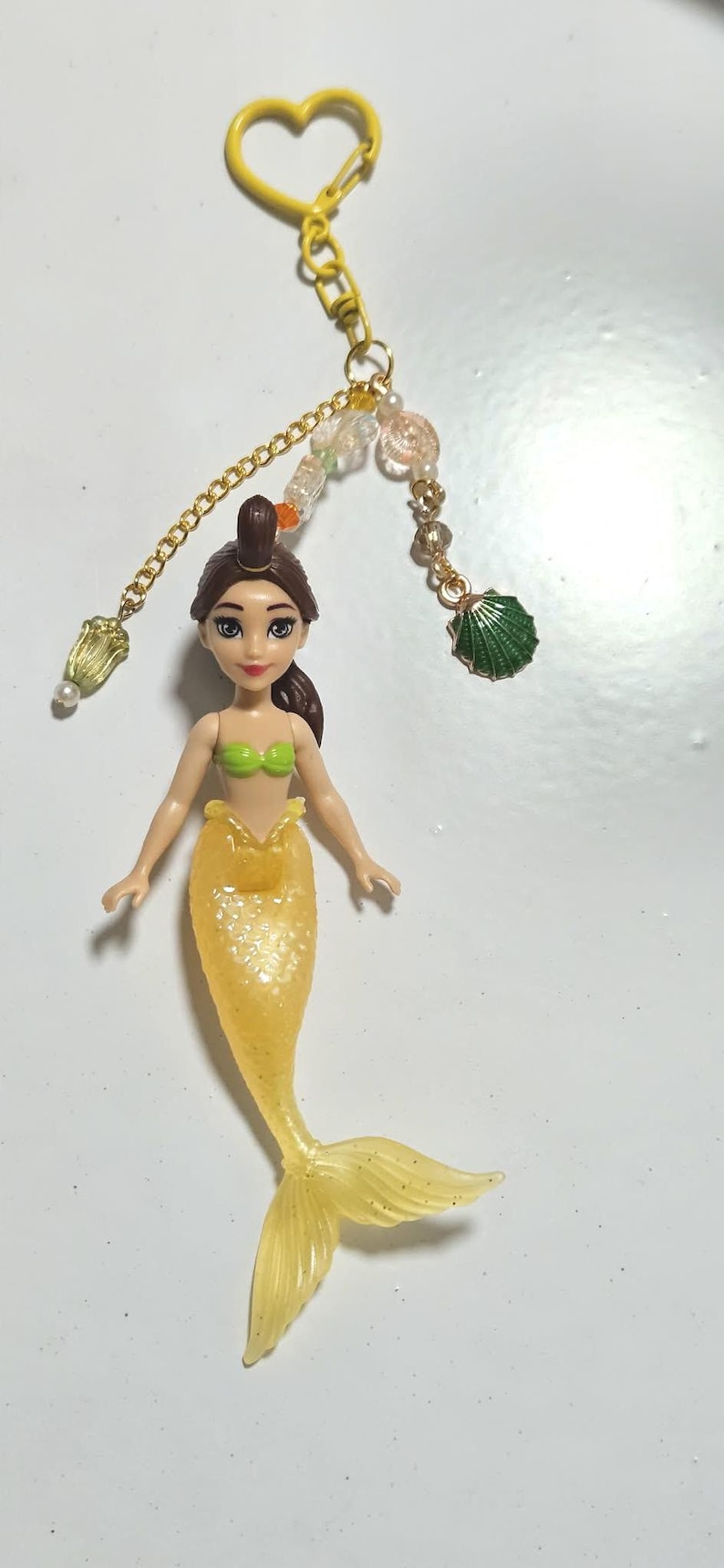 May include: A mermaid keychain featuring a yellow tail and green top. The doll has brown hair and is attached to a gold chain with decorative beads and a yellow heart-shaped clasp. A green seashell charm is also included.
