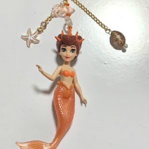 May include: A mermaid keychain with an orange tail and top. The keychain has a gold chain with a starfish charm, a cloud charm, and a decorative bead. The mermaid has brown hair and is holding her arms out.