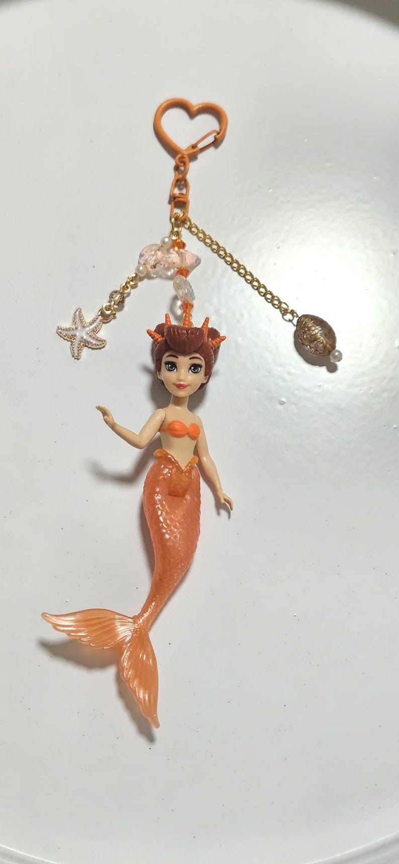 May include: A mermaid keychain with an orange tail and top. The keychain has a gold chain with a starfish charm, a cloud charm, and a decorative bead. The mermaid has brown hair and is holding her arms out.