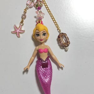 May include: A pink mermaid doll keychain with a gold chain, pink heart-shaped clasp, and decorative beads. The mermaid has a pink tail, a pink top, and blonde hair. A pink starfish charm is also attached.