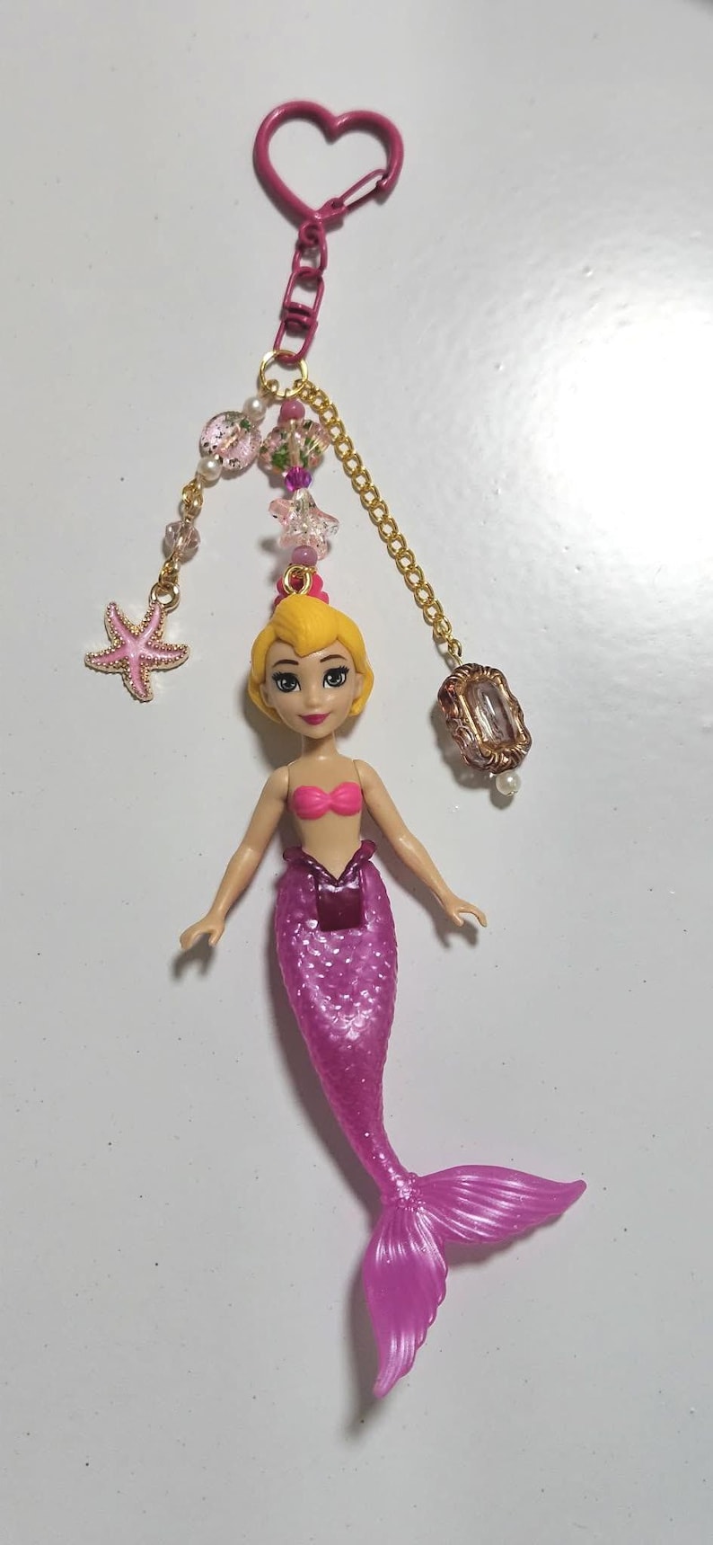 May include: A pink mermaid doll keychain with a gold chain, pink heart-shaped clasp, and decorative beads. The mermaid has a pink tail, a pink top, and blonde hair. A pink starfish charm is also attached.