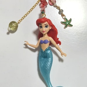 May include: A keychain featuring a mermaid doll with red hair, a blue tail, and a purple top. The keychain has a gold chain with decorative beads and a green starfish charm. The clasp is purple.