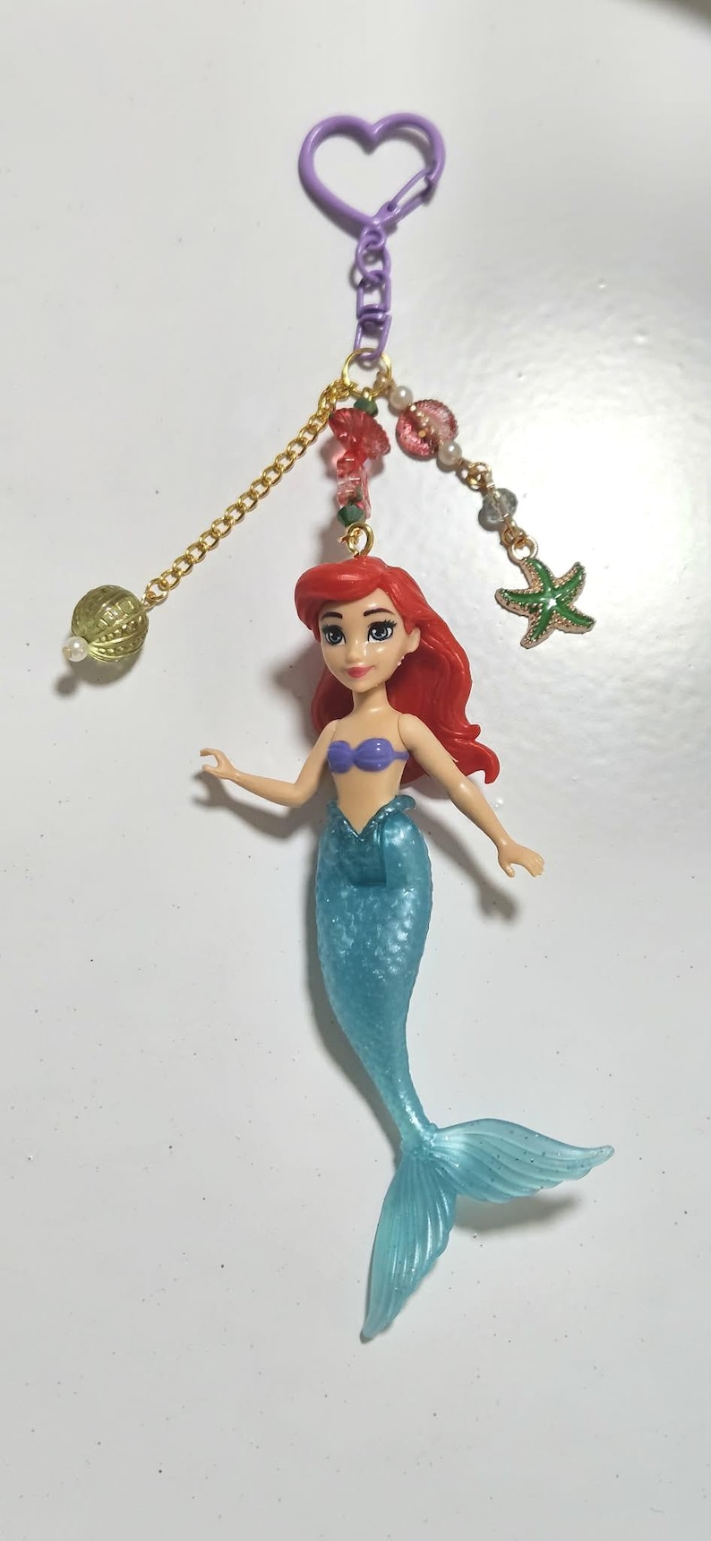 May include: A keychain featuring a mermaid doll with red hair, a blue tail, and a purple top. The keychain has a gold chain with decorative beads and a green starfish charm. The clasp is purple.