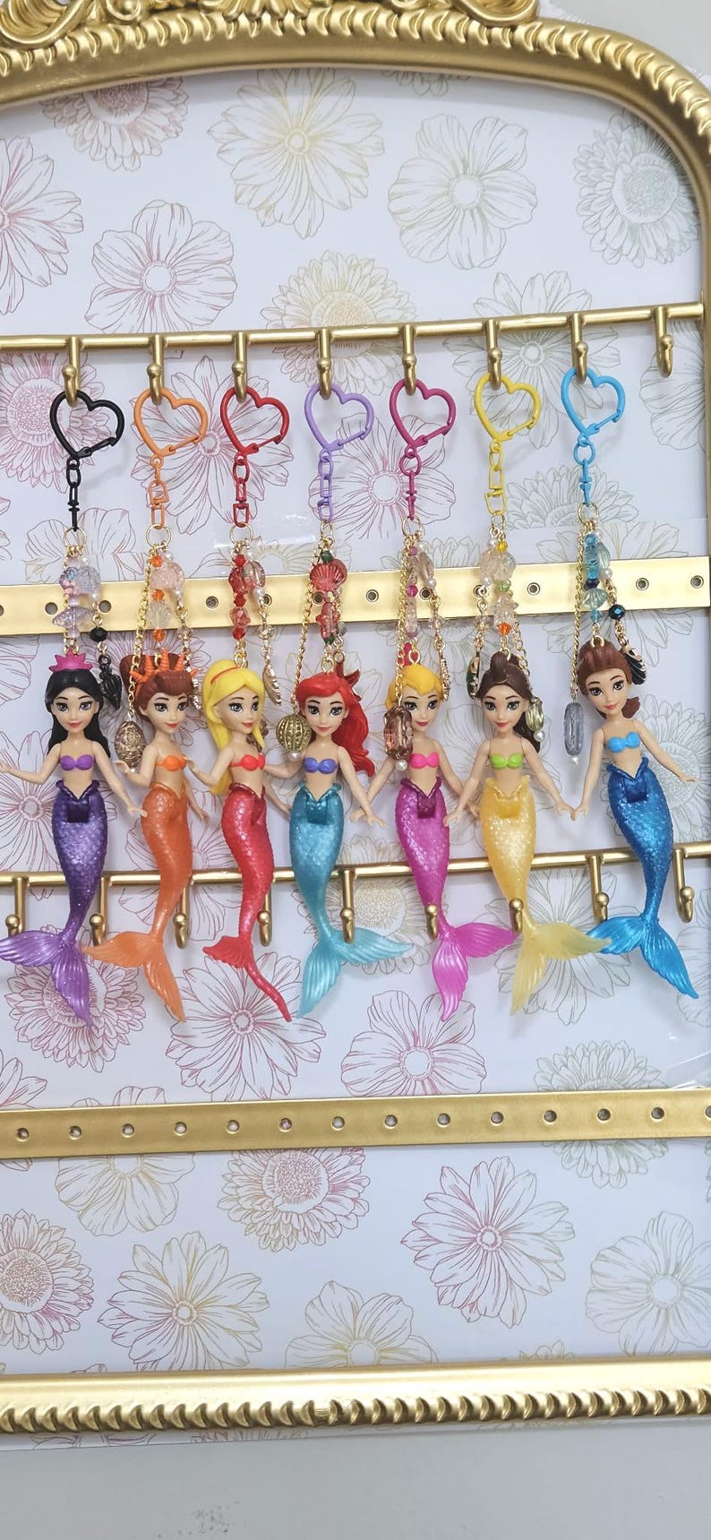May include: A collection of mermaid keychains with colorful tails and tops, each attached to a heart-shaped keyring. The mermaids are displayed on a gold-framed rack with a floral background.