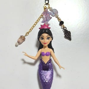 May include: A mermaid keychain featuring a purple tail and a purple top. The doll has black hair and a pink crown. The keychain includes a heart-shaped clasp, gold chain, and decorative beads. The mermaid is approximately 5 inches tall.