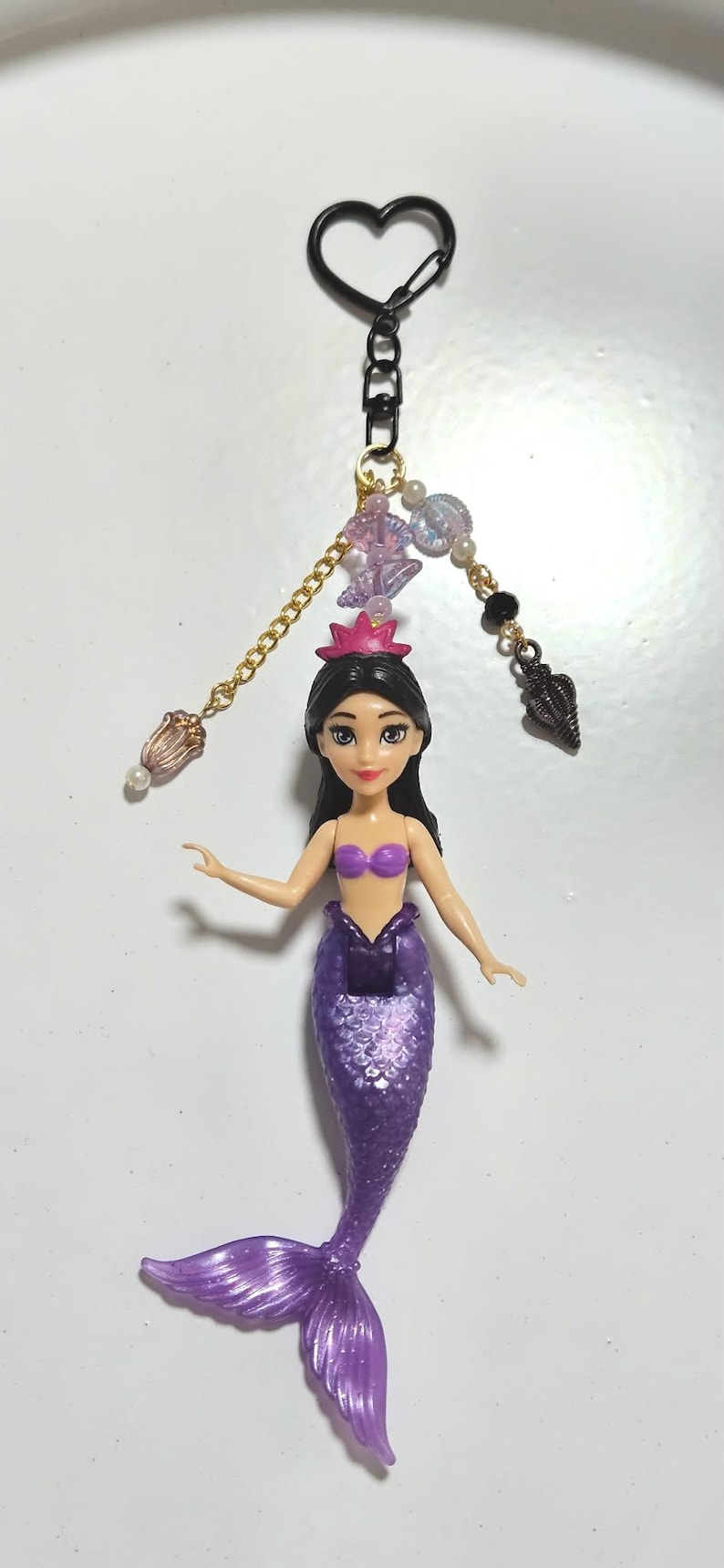 May include: A mermaid keychain featuring a purple tail and a purple top. The doll has black hair and a pink crown. The keychain includes a heart-shaped clasp, gold chain, and decorative beads. The mermaid is approximately 5 inches tall.