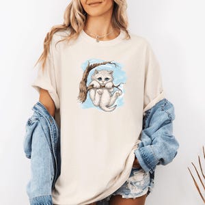 Pode incluir: Camiseta creme com uma ilustração em aquarela de um gatinho branco pendurado em um galho de árvore. O gatinho tem olhos azuis e está em um fundo azul claro. A camiseta tem um caimento folgado.