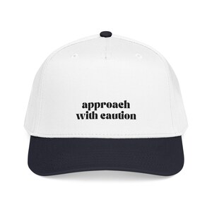 Puede incluir: Gorra de béisbol blanca con visera azul marino y botón negro. La frase "approach with caution" está impresa en letras negras en negrita en la parte delantera de la gorra.