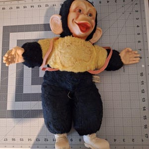May include: Vintage monkey doll with a smiling face, large ears, and open mouth. The doll has a black head and arms, a yellow torso, and black pants. It wears white shoes and has a pink strap across the chest. The doll is lying on a gray cutting mat.