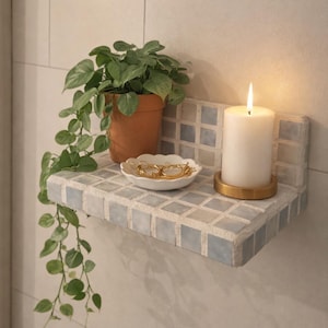 May include: A small, tiled shelf with a potted plant, a white candle in a gold holder, and a small white dish with gold jewelry. The shelf is mounted on a light-colored tiled wall.