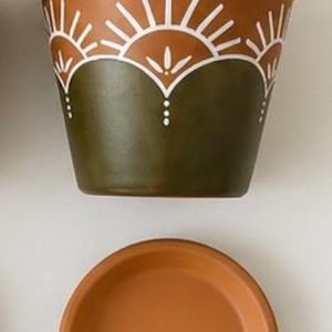 May include: A terracotta-colored flower pot with a matching saucer. The pot is painted with a white sunburst design on the top and a dark green base. The saucer is round and has a slightly raised edge.