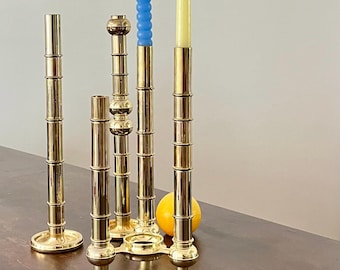 May include: A collection of brass candle holders in varying heights and designs. Two holders have blue and yellow taper candles. The candle holders are on a dark brown surface with a yellow orange fruit.