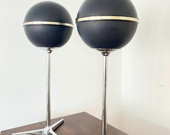 May include: Two vintage black globe lamps with silver-colored trim and a chrome base. The lamps have a textured surface and are on a dark wooden table. The lamps are a unique lighting fixture.