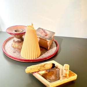 May include: A collection of spiritual items, including a beige candle, a small wooden box, a ceramic incense holder, and a palo santo stick with ash. The items are arranged on a decorative plate with a red rim.