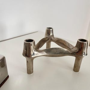 May include: A silver three-armed candle holder with a modern, abstract design. The holder has a smooth, polished finish and is made of metal.