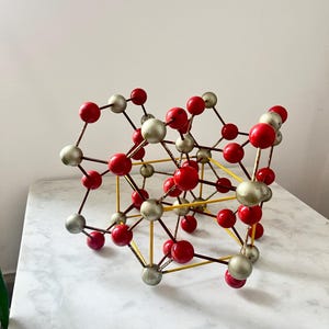 Pode incluir: Modelo tridimensional de uma estrutura molecular. Apresenta esferas vermelhas e prateadas conectadas por hastes marrons e suportes amarelos, criando um design geométrico complexo. As esferas estão dispostas em um padrão de treliça.