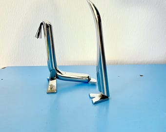 Puede incluir: Dos esculturas de metal plateado de pájaros estilizados. Los pájaros están de pie sobre una superficie azul. El pájaro de la izquierda tiene un pequeño borla negra en la cabeza.