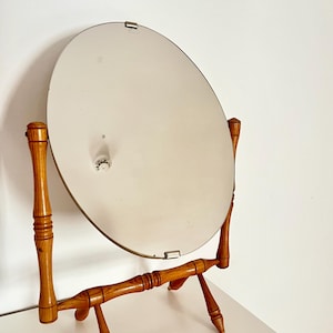 May include: Oval vanity mirror with a light brown wooden frame and legs. The mirror is supported by two vertical posts and a horizontal bar, with a small, round base. The mirror is tilted slightly back.