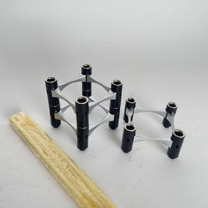 Könnte beinhalten: Set aus drei Kerzenhaltern mit modernem Design. Die Kerzenhalter sind schwarz und silberfarben, mit einer geometrischen Form. Eine Packung langer, dünner Kerzen ist ebenfalls enthalten.