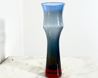 Könnte beinhalten: Eine elegante Glasvase mit Farbverlauf. Der obere Teil ist hellblau, geht in Grau über, dann in Dunkelblau und endet mit roten Streifen am Boden. Die Vase hat einen ausgestellten oberen Teil und einen schlanken, länglichen Körper.