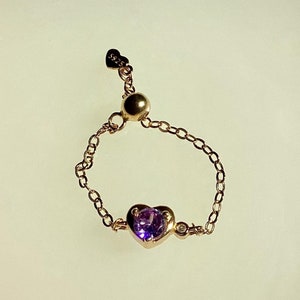 以下が含まれることがあります： 紫色のハート型の宝石と小さな金色のビーズが付いた、繊細な金のチェーンリング。リングは白い背景の上に置かれています。