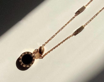 May include: A rose gold necklace with a round pendant featuring Roman numerals and a black center. The necklace has two small rectangular charms.