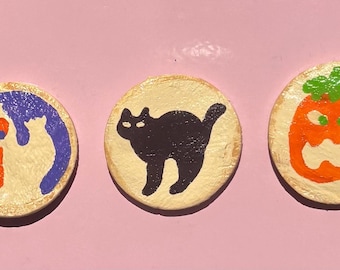 May include: Three round Halloween-themed decorations. One features a ghost with a blue outline and orange mouth, the second a black cat, and the third an orange jack-o'-lantern with a green stem. All are on a light beige background.