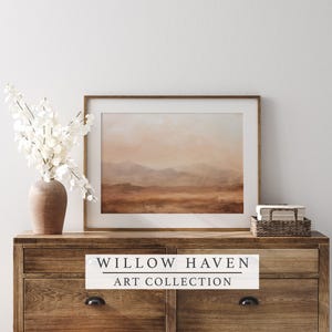 May include: A framed landscape art print with a warm, neutral color palette. The artwork depicts a mountain range and a hazy sky. The print is displayed on a wooden dresser, alongside a vase of white flowers and a small woven basket. The text "WILLOW HAVEN ART COLLECTION" is displayed on the dresser.